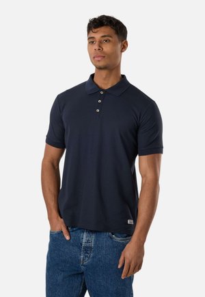 Mand stående iført en mørke marineblå polo med korte ærmer og knapper samt blå jeans, hænderne afslappet ved siderne mod ensartet baggrund.
