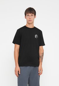 Sort bomulds T-shirt med korte ærmer, rund halsudskæring, og et hvidt logo trykt på venstre bryst. Modellen har grå shorts på.
