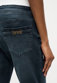 Κοντινό πλάνο στην πίσω τσέπη σκούρων τζιν με χρυσό λογότυπο "Versace Jeans Couture", εμφανίζοντας μέρος του χεριού και του γοφού ενός ατόμου.