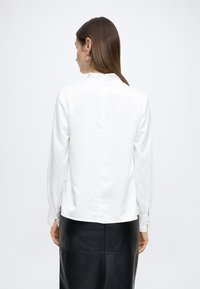 Bluză albă cu mâneci lungi ușor bufante, guler înalt și fermoar ascuns în spate. Material neted, lucios, cu croială ajustată.