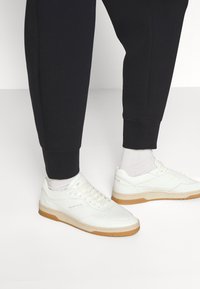 Białe skórzane sneakersy z perforowanym noskiem, beżowymi akcentami i gumową podeszwą, w zestawieniu z czarnymi joggerami i jasnymi skarpetami.