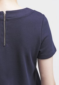 Top en tricot bleu marine à manches courtes et col rond. Présente une fermeture éclair visible à l'arrière et un design en tissu texturé.