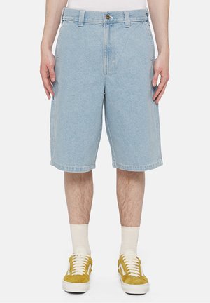 Homme portant un short en denim bleu clair, des chaussettes blanches et des baskets jaune moutarde sur un fond blanc.