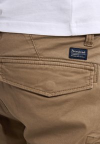 Pantalones caqui con una textura suave, que cuentan con un bolsillo trasero y una etiqueta de marca azul que dice "Petrol Ind." Costuras visibles a lo largo de las costuras.