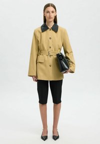 Giacca impermeabile khaki con colletto nero, cintura in vita e tasche laterali; abbinata a pantaloni shorts neri su misura e décolleté neri a punta.