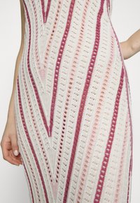 Vestido de punto con un diseño ajustado en crema, que presenta rayas rosas y fucsias y patrones de agujeros abiertos. Texturizado con un ligero brillo.