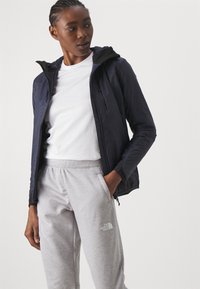 Marine letvægtsjakke med hætte, sort foer og sidelommer, parret med grå sweatpants med en logo på venstre lår.