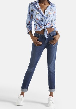 Chemise bleue nouée à l'avant avec imprimés graphiques, associée à un jean en denim foncé, ourlets retroussés et baskets blanches. Style décontracté et ajusté.