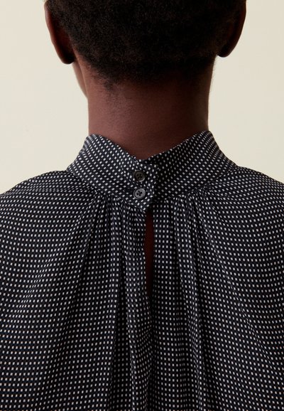 Blusa con estampado en blanco y negro que presenta un cuello alto, pequeños botones en el cuello y un detalle de pliegues en la parte posterior. Textura de tejido suave.