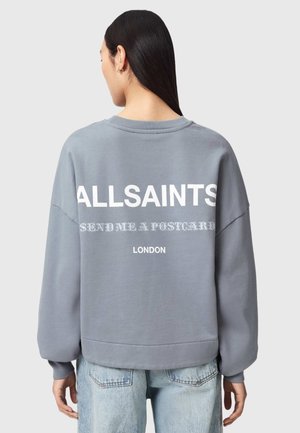Frau mit langen dunklen Haaren, die ein graues Sweatshirt mit dem Aufdruck „ALLSAINTS SEND ME A POSTCARD LONDON“ auf der Rückseite trägt, kombiniert mit hellblauen Jeans.