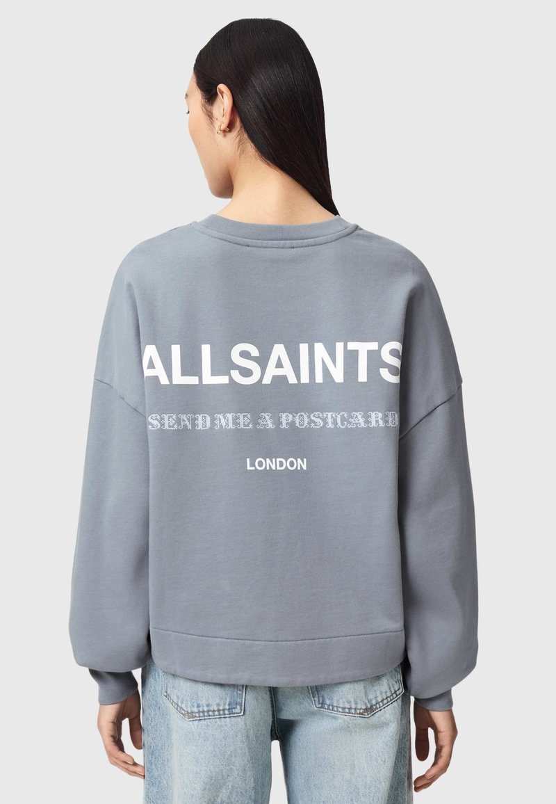 Woman with long dark hair wearing a gray sweatshirt with “ALLSAINTS SEND ME A POSTCARD LONDON” printed on the back, paired with light blue jeans.