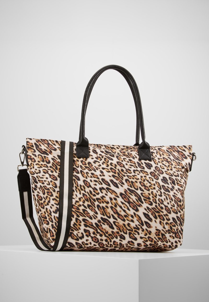 Grand sac cabas en imprimé léopard avec des poignées en cuir noir et une bandoulière amovible rayée noir et blanc reposant sur une surface blanche.