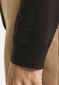 Schwarzer gestrickter Pulloverbund mit geripptem Textur und dezenter Logodetail. Tanfarbener Stoff im Hintergrund sichtbar, der den Kontrast hervorhebt.