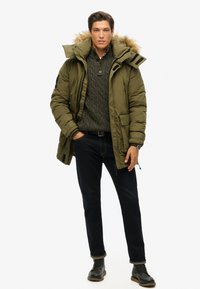 Oliven grøn pufferjakke med pelsforing på huen, parret med en mørk striktrøje og mørke denimjeans, iført sorte snørebåndsstøvler.