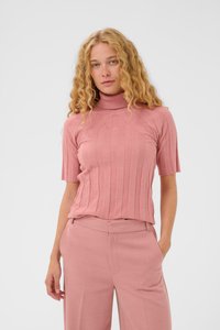 Kvinde med krøllet blondt hår iført en rillede pink kortærmet turtleneck sweater og matchende brede bukser, stående med den ene hånd i lommen.