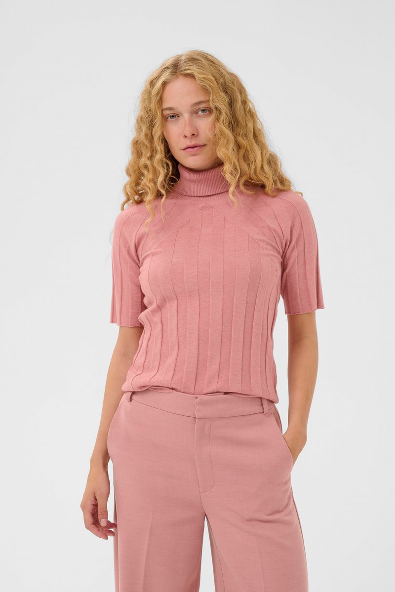 Kvinde med krøllet blondt hår iført en rillede pink kortærmet turtleneck sweater og matchende brede bukser, stående med den ene hånd i lommen.