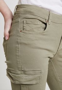 Pantalones cargo verde oliva claro hechos de una tela suave, con múltiples bolsillos, costuras visibles y una cintura ajustada con un pequeño detalle de botón.