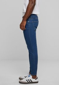 Jeans ajustados azules con cinco bolsillos, hechos de tela vaquera. Presentan detalles de costura y un ligero desgaste. Llevados con zapatillas blancas y negras.