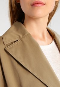 Béžový trenčkot s ostrým límcem, viditelnými švy a hladkým povrchem. Nosený přes světle zbarvený oděv.