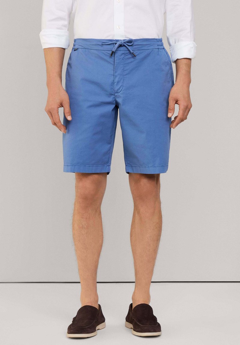 Pantaloni corti in cotone blu con vita elastica e coulisse. Presentano tasche laterali e una lunghezza a metà coscia. Indossati con scarpe scure senza lacci.