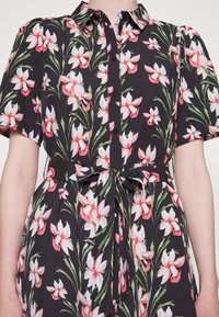 Vestido preto florido com flores rosas e brancas, mangas curtas, frente com botões e uma cintura amarrada para uma silhueta ajustada.