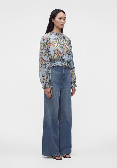 Femme debout en jean bleu à jambes larges, blouse à fleurs bleu clair à manches longues et sandales noires, sur fond blanc uni.