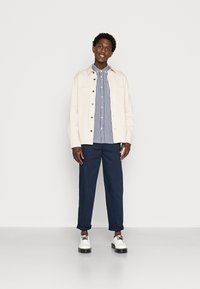 Chaqueta beige clara, camisa a cuadros en azul y blanco, pantalones azul marino y zapatos blancos con suelas negras. Texturas suaves y ajuste entallado.