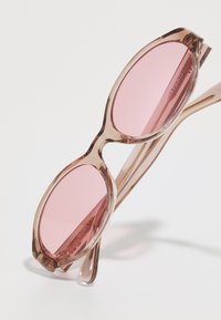 Lunettes de soleil rose clair avec une monture transparente, des verres ovales et des branches élégantes, présentant une finition brillante et des bords arrondis.