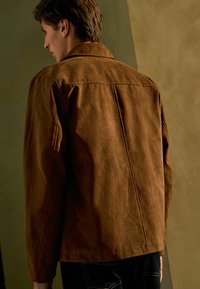 Chaqueta de ante marrón con un collar clásico, corte relajado y detalles de costura visibles. Llevada sobre pantalones oscuros, vista desde atrás.