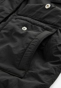 Schwarzer, leichter Stoff mit strukturierter Oberfläche. Verfügt über eine große Tasche mit Druckknopf, glatten Kanten und minimalen Nahtdetails.