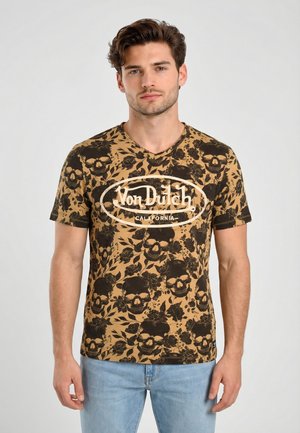 Hombre joven vistiendo una camiseta marrón de Von Dutch con un estampado negro de flores y calaveras, y jeans azul claro, posando frente a un fondo liso.