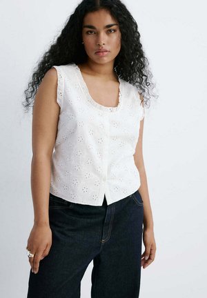 Frau mit langen lockigen schwarzen Haaren, die eine ärmellose weiße Bluse mit Lochmuster und dunkelblaue Jeans trägt, vor einem schlichten Hintergrund stehend.