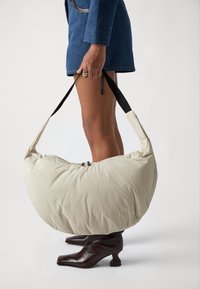 Bolso acolchado beige con una forma grande y curva, correas negras y cierre de cremallera. Modelo lleva botas de tacón marrón oscuro y una chaqueta azul.
