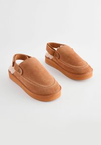 Brune ruskinds slip-on sko med en rund tå, hvid syning og en blød indvendig foring. Har et justerbart bånd ved hælen.