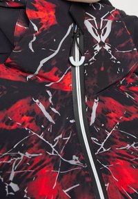 Hooded jacka i svart med röd och vit abstrakt mönster, med frontdragkedja, snörning och lätt, texturerad tyg.