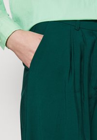 Grön långärmad skjorta som är kombinerad med mörkgröna veckade byxor, som har en sidoficka och en mjuk tygkvalitet.
