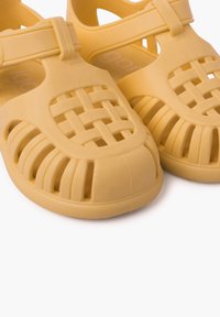 Gelbe Plastiksandalen mit einem geflochtenen Oberdesign, offenen Seiten und einer dicken Sohle. Enthalten einen verstellbaren Riemen über dem Fußrücken.