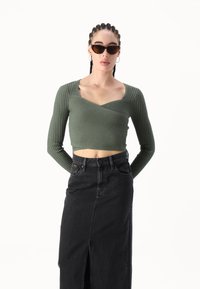 Olivengrønn, ribbet crop top med lange ermer, med en V-halsdesign. Kombinert med et høytsittende svart denimskjørt, med spalte detalj synlig.