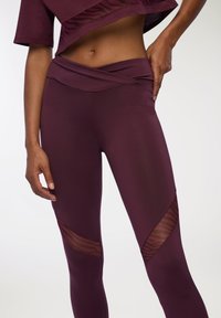 Leggings bordeaux con vita alta, caratterizzati da pannelli in rete con un sottile motivo a righe sulle cosce esterne. Tessuto liscio e elastico.