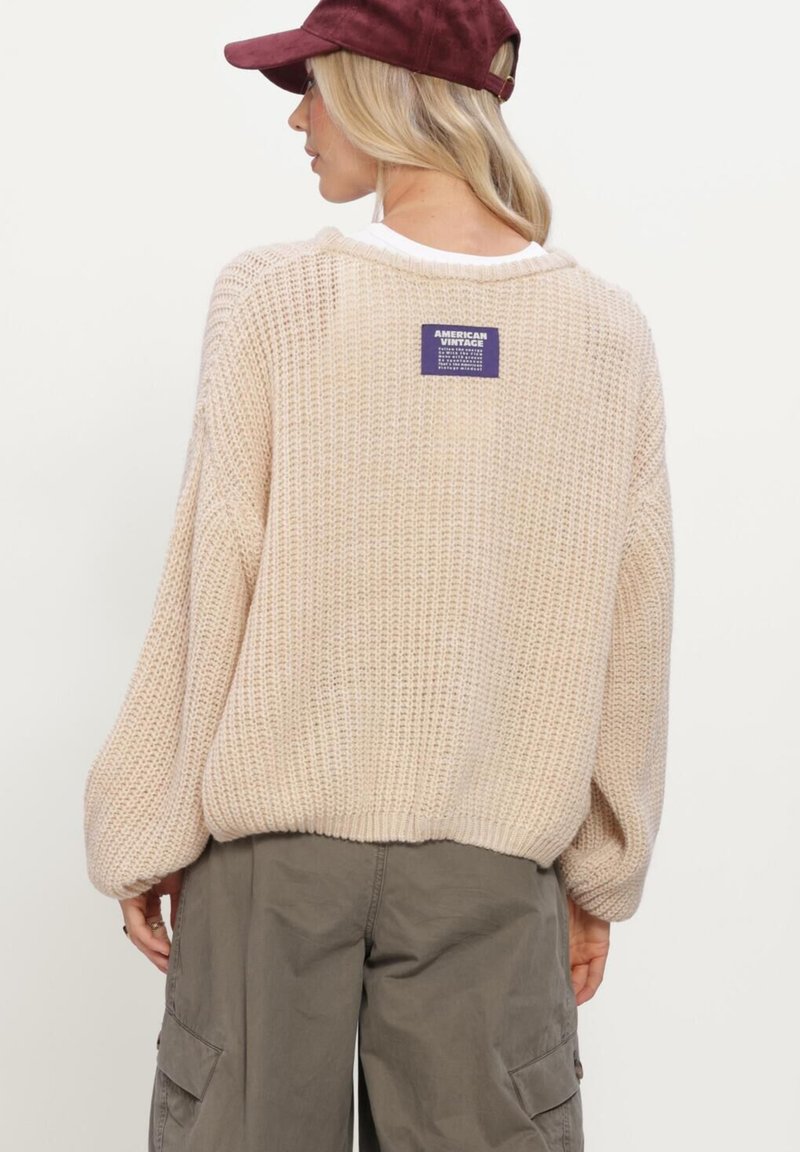 Blonde woman wearing a beige knit sweater with a label on the upper back, gray cargo pants, and a burgundy cap, facing away from the camera.