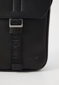 Sort læder messenger taske med tekstureret overflade, sølv spænde hardware og prægede "CK" logo foran. Minimalistisk design.