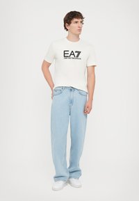 White T-shirt with black EA7 logo, paired with light blue, loose-fitting jeans. White sneakers complete the casual look.