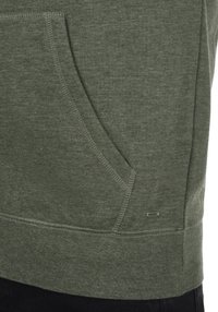 Groene pullover met een voorzak, ribbelrand en naden. Gemaakt van zachte stof, met een klein metalen logoaccent aan de zijkant.