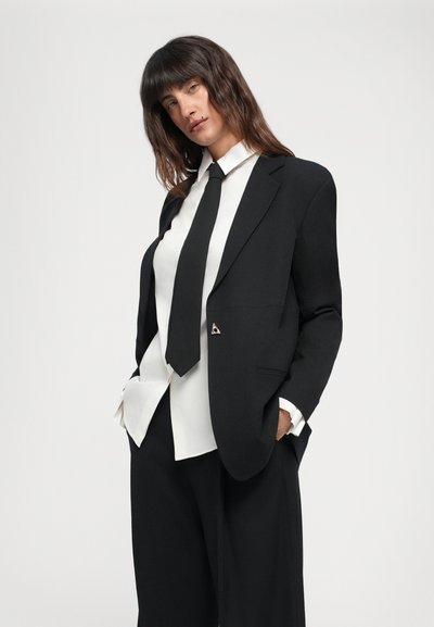 Black tailored blazer with sharp lapels, paired with a white shirt and black tie. Features silver triangle hardware and loose-fitting black trousers.