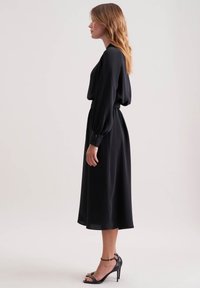 Donna che indossa un vestito midi nero a maniche lunghe con vita cintrata e sandali neri con cinturino alla caviglia e tacco alto, in piedi di profilo.