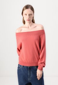 Pull tricot corail à épaules dénudées avec des poignets et un ourlet côtelés, présentant une texture douce et une coupe décontractée, associé à un jean en denim bleu ample.