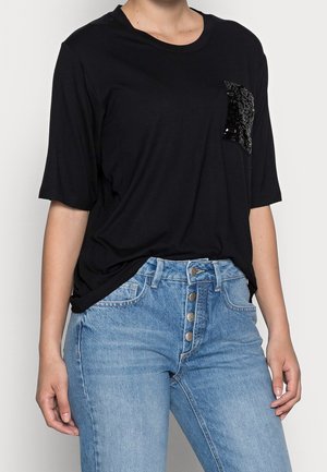 Mujer con una camiseta negra de manga corta con un bolsillo en el pecho adornado con lentejuelas, combinada con jeans azul de cintura alta y cierre de botones.