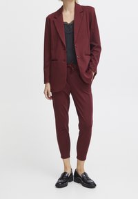 Femme portant un blazer bordeaux et un pantalon à cordon assorti, avec un haut noir orné de dentelle et des mocassins noirs, debout sur un fond blanc.