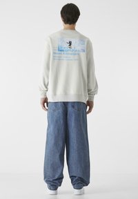 Világosszürke pulóver, hátul grafikai részlettel, amely a Kilimandzsáró hegyet, a magassági adatokat és egy mászó illusztrációt ábrázol. Laid-back kék farmernadrággal viselve.