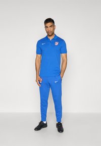 Blå atletisk polo og joggingbukser med Nike-logo og holdemblem. Poloen har krave; bukserne har taperede manchetter. Bæres med sorte sportssko.